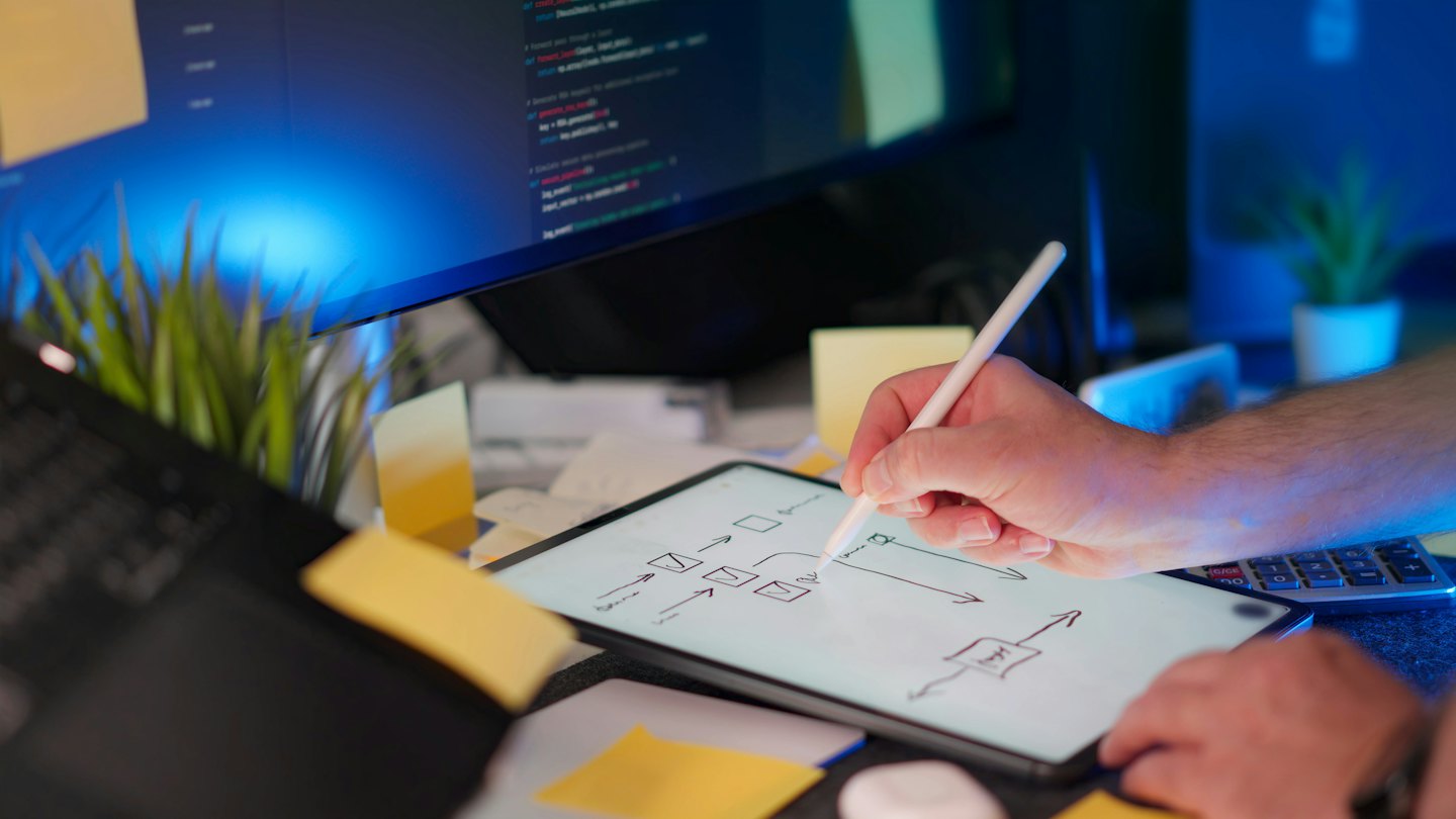 Hand using stylus to draw flowchart on tablet with computer monitor.